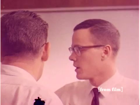 This still from the Bell System film 'Preventing Data Transmission Errors' features two engineers engaged in conversation. The individuals are dressed in professional attire typical of the late 1960s, wearing glasses and showing focused expressions, indicating a discussion possibly related to computer technology or data transmission. The background suggests a lab or classroom setting, typical for technical training or presentations. The color tones are muted, consistent with the film's age, and there are visible defects characteristic of older film stock.