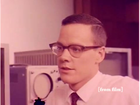 The still from the Bell System film 'Preventing Data Transmission Errors' features a young man with glasses, dressed in a button-up shirt and tie, likely an engineer or technician. He appears engaged in conversation, possibly discussing topics related to data transmission and technology. In the background, there are computers and oscilloscope equipment, highlighting the film's focus on telecommunications and error prevention methods in data transmission. The overall aesthetic reflects the 1960s style of both attire and technology, presenting a glimpse into the era's innovation in computing.