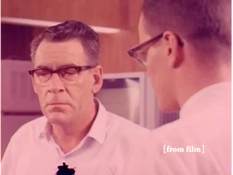 The still shows two men in a setting likely related to a technology or engineering environment, presumably from a Bell System film. One person appears to be focused or contemplative, while the other seems engaged in conversation, possibly discussing data transmission or related topics. The color is muted, reflecting the film's age and silent nature. There seems to be a lab defect, indicated by an anomaly in the image. This still encapsulates the era's emphasis on technology and communication.