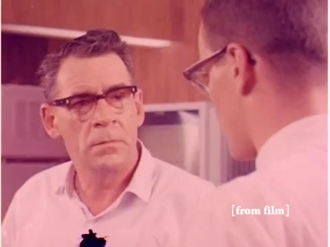 The still from 'Bell System - Preventing Data Transmission Errors' depicts two engineers engaged in a serious conversation. One individual, wearing glasses and a light-colored shirt, displays a concerned expression, while the other, also in glasses and a white shirt, appears to be explaining or discussing something. The background features a subtle, professional setting, likely within a lab or office environment. The film, a silent 16mm, captures the essence of the technological discussion typical of the era, although this still shows signs of a lab defect, affecting the visual quality.