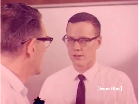 The still captures a moment from the Bell System film 'Preventing Data Transmission Errors,' produced in 1967. It features two men wearing glasses engaged in a discussion, possibly about technical topics related to data transmission. One man is positioned to the left, while the other is facing him. The background appears to include a wall, suggesting a laboratory or office setting. Overall, the image reflects a period of early computing and telecommunications development, aligned with the film's focus on technology and error prevention.