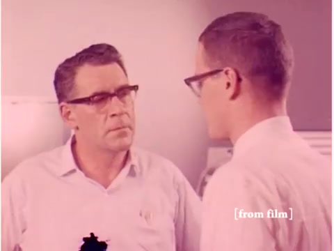 The still from 'Bell System - Preventing Data Transmission Errors' features a conversation between two men, likely engineers, in a lab environment. Their attire, including glasses, suggests a professional setting typical of the late 1960s. The background indicates a technical workspace, possibly with equipment relevant to data transmission. The film has a color defect, giving it an unusual hue. The focus appears to be on the discussion about data transmission, reflecting the technological advancements of that era.
