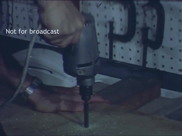 The still features a close-up of a hand holding a drill, actively working on a piece of wood. In the background, a white telephone model is visible, suggesting a connection to the Bell Trimline Extension theme. The setting appears to be a workshop, indicated by the pegboard and tools visible in the frame. The image is part of a commercial, aimed at promoting the usefulness of the telephone in home or DIY projects. The 'Not for broadcast' label indicates that this still is not intended for public airing.