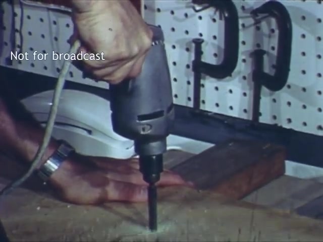 The still from the 'Bell Trimline Extension - C&P telephone commercial' features a close-up of a hand gripping a power drill, poised above a piece of wood. In the background, a white Bell Trimline telephone is visible on a work surface. The setting suggests a DIY or home improvement context, highlighted by the organized pegboard showing various tools and clamps. The focus on the drilling action implies a demonstration or instructional element, likely promoting the ease of installation associated with the telephone extension.