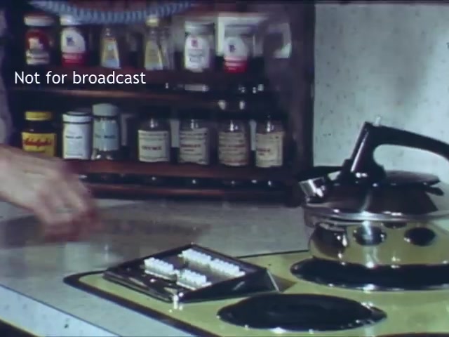 The still from the 'Bell Trimline Extension - C&P telephone commercial' features a kitchen setting with a countertop and various household items. Notably, there's a kettle on the stove, and a tray with what appears to be telephone accessories. Behind, a shelf is stocked with jars, suggesting a homey, domestic atmosphere. The scene conveys a sense of everyday life and utility, complementing the theme of the commercial.