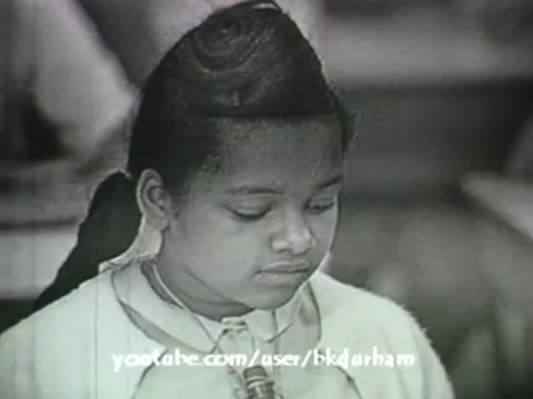 The still from '6th Grade Linguistics (1966)' showcases a young girl with a thoughtful expression, focused on her work. The monochrome aesthetic and soft contrast give it a vintage feel reminiscent of a kinescope. The classroom setting hints at an educational context, aligning with its purpose as a companion to a textbook. The image captures a moment of concentration, reflecting the learning experience of the time.