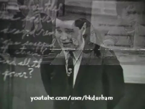 The still from '6th Grade Linguistics (1966)' captures a classroom scene featuring a young boy in a suit, likely delivering a speech or presentation. The image exhibits a vintage aesthetic, resembling a kinescope, with a slightly blurred quality and overlay of handwritten notes. This educational film integrates visual elements to engage sixth graders in linguistic concepts, reflecting mid-20th century teaching methods. The background features a chalkboard with sketches, enhancing the academic context.