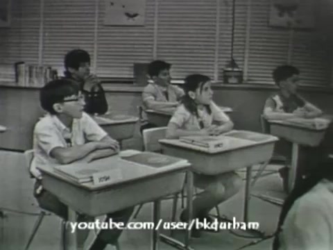 The still from '6th Grade Linguistics (1966)' depicts a classroom scene featuring several students attentively facing a teacher or presentation. The black-and-white image has a vintage quality, reminiscent of kinescopes, with visible grain. The students, seated at their desks, exhibit a mix of curiosity and concentration, reflecting a typical educational setting of the era. The environment is simple, with basic classroom decor and learning materials visible in the background, evoking the educational practices of the 1960s.