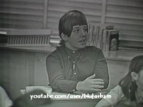 The still from '6th Grade Linguistics (1966)' features a classroom scene with a focus on a boy with a distinctive hairstyle, wearing a buttoned-up shirt. He appears engaged in conversation, suggesting a lively atmosphere typical of an educational setting. The monochromatic aesthetic and slight grain give it a vintage feel reminiscent of kinescopes, emphasizing the film's historical context as a teaching aid from the 60s. The background includes shelves with books, reinforcing the educational theme.
