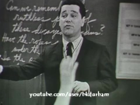 The still from '6th Grade Linguistics' (1966) features a male teacher addressing a classroom. He appears engaged, possibly asking a question or encouraging participation, as a student's hand is raised in response. The background includes handwritten notes on a blackboard, indicative of the educational setting. The overall look has a vintage quality, reminiscent of kinescope film style, capturing the era's educational aesthetics.