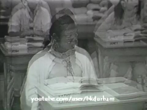 The still from '6th Grade Linguistics (1966)' depicts a classroom setting reminiscent of a kinescope with a vintage film quality. It features a student seated at a desk, surrounded by stacks of books. The image has a slightly faded appearance, characteristic of early educational films, and conveys a sense of nostalgia. The blurred, echo-like effect adds to its age, emphasizing the film's historical context as a tool for teaching linguistics in schools during that era.