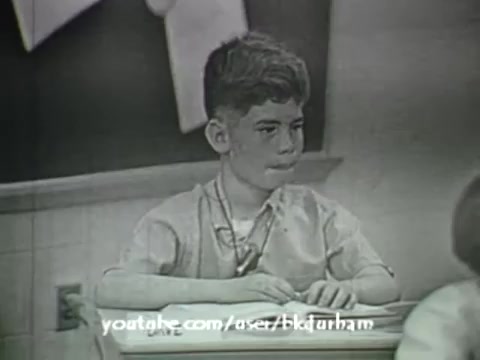 The still from '6th Grade Linguistics (1966)' features a young boy sitting at a classroom desk, seemingly engaged in thought or concentration. The monochrome aesthetic, reminiscent of kinescope recordings, adds a nostalgic quality to the image. The background includes elements typical of a classroom setting, such as a chalkboard. This educational film aimed to complement a textbook, reflecting mid-20th century pedagogical approaches.