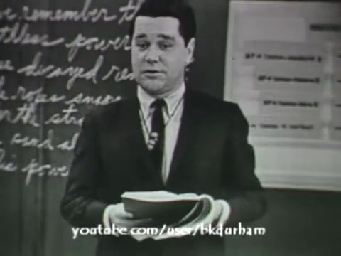 The still from '6th Grade Linguistics' (1966) captures a classroom scene in black and white, reminiscent of kinescope films. A male instructor stands at the front, holding several books. Behind him is a chalkboard with handwritten notes, suggesting an educational context. The overall composition conveys a mid-20th-century teaching style, reflective of the era's approach to linguistic education.