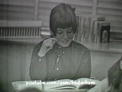The still from '6th Grade Linguistics' (1966) features a young student with a bobbed haircut, smiling as she interacts with a textbook. She appears engaged, possibly reflecting on the material. The black-and-white cinematography gives it an old-school educational feel, reminiscent of kinescope recordings, indicating its vintage nature. The classroom setting, with other students partially visible, enhances the educational context.