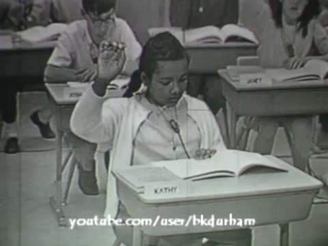 The still from '6th Grade Linguistics' features a classroom scene with a young girl raising her hand, presumably to answer a question or participate in discussion. She sits at a desk with an open textbook, while classmates are visible in the background, also engaged in the lesson. The monochrome aesthetic gives it a vintage feel, reminiscent of kinescope broadcasts. This educational film aims to complement the instructional material and highlights an interactive classroom dynamic.