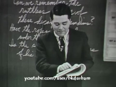 The still from '6th Grade Linguistics' (1966) features a male teacher in front of a chalkboard. He appears to be engaged with his textbook, possibly discussing linguistic concepts. The chalkboard displays handwritten notes, adding to the educational context of the film. The visual quality suggests a vintage aesthetic, reminiscent of kinescope recordings, highlighting the era's film techniques for educational purposes.