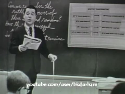 The still from '6th Grade Linguistics' (1966) presents a classroom scene characterized by a young male teacher standing at the front, holding a paper and engaging with students. Behind him, a chalkboard features illustrative notes, while a chart with categorized information is displayed on the wall. The overall monochromatic aesthetic gives it a vintage, kinescope-like appearance, emphasizing its educational context and era.