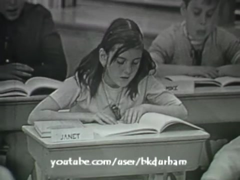 The still features a classroom scene from the 1966 educational film '6th Grade Linguistics'. A young girl, identified as Janet, is focused intently on her textbook, surrounded by classmates. The monochromatic image carries a vintage aesthetic reminiscent of kinescopes, highlighting the educational atmosphere of the time. The setup suggests a structured learning environment typical of the era, emphasizing engagement with linguistics.
