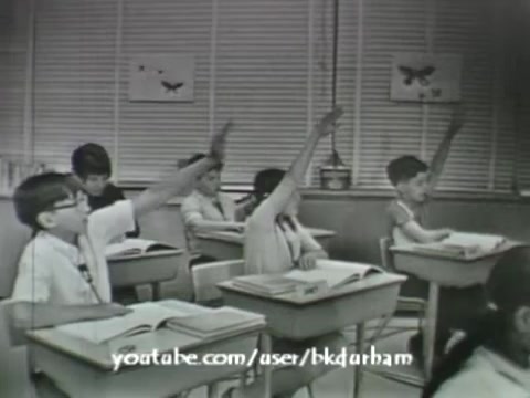 The still from '6th Grade Linguistics' (1966) depicts a classroom scene with several students raising their hands, suggesting an engaged atmosphere of learning. The monochrome aesthetic gives it a vintage feel reminiscent of kinescopes, emphasizing an era of educational film. The students are seated at desks with textbooks open, indicating an interactive lesson. The background features simple classroom decor, typical of the 1960s educational settings.