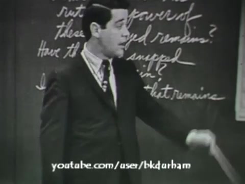 The still from '6th Grade Linguistics' (1966) features a teacher in a suit standing in front of a chalkboard, engaging with students. The chalkboard displays handwritten text, suggesting a focus on language or grammar. The film's black-and-white quality and directorial style resemble that of a kinescope, emphasizing its educational purpose. The teacher appears animated, likely discussing concepts related to linguistics, with a pointer in hand.