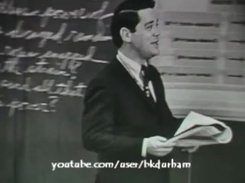 The still from '6th Grade Linguistics' (1966) features a male presenter in a suit, standing in front of a chalkboard filled with handwritten notes. He appears engaged and is holding papers, likely teaching a lesson on linguistics. The overall aesthetic resembles a kinescope, emphasizing the educational context of the film. The image's quality and framing capture a classic mid-20th-century classroom setting.