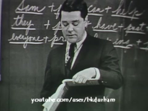 The still features a male educator in a suit, standing in front of a chalkboard filled with handwritten notes, likely presenting a lesson on grammar or linguistics. The image captures a vintage aesthetic reminiscent of kinescopes, with a monochromatic palette that underscores its educational purpose from the 1960s. The instructor appears engaged, holding a book or teaching aid while addressing the camera or class. The overall composition reflects the formal educational style of the era.