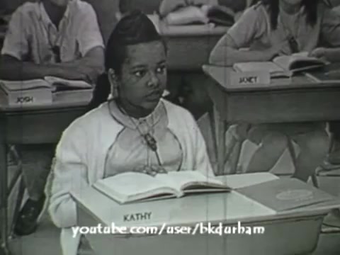 The still from '6th Grade Linguistics (1966)' features a young girl, Kathy, sitting at her desk in a classroom, focused on an open textbook. The black-and-white imagery resembles a kinescope, adding a retro educational aesthetic. In the background, other students are partially visible, contributing to the classroom setting. The overall composition emphasizes a traditional learning environment typical of the era.