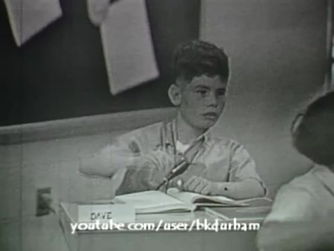 The still from '6th Grade Linguistics' (1966) features a young boy named David sitting at a classroom desk, looking intently at a book while raising his hand, possibly to answer a question. The monochrome quality and overall aesthetic evoke a vintage, kinescope-like feel, typical of educational films from that era. The setting suggests a structured learning environment focused on linguistics, engaging students in active participation.