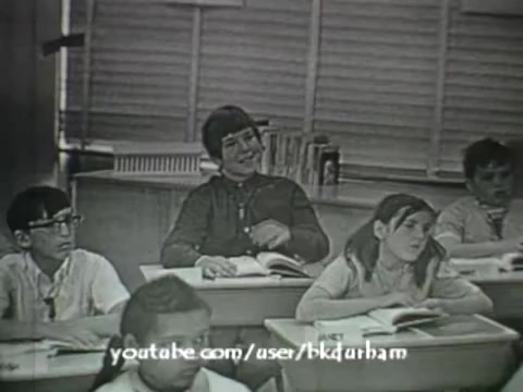 The still from '6th Grade Linguistics (1966)' features a classroom scene with several students engaged in a lesson. The film has a vintage quality, akin to a kinescope, characterized by its monochrome palette. The students display a range of expressions, with one boy smiling and another girl appearing more disengaged. The educational context is evident through the textbooks in front of them and the classroom setting, which contains wooden desks and a window with blinds. The overall ambiance suggests a moment of interactive learning typical of mid-20th century educational films.