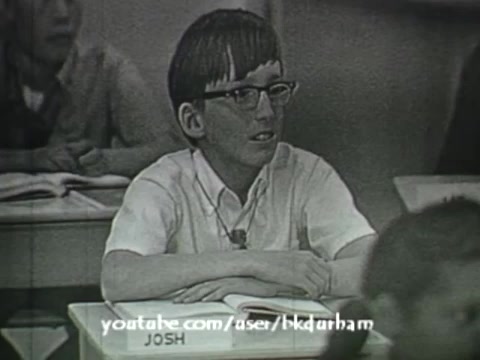 The still from '6th Grade Linguistics' (1966) presents a black-and-white scene reminiscent of a kinescope. It features a young boy named Josh sitting at a school desk, with books and materials in front of him. His focused expression suggests engagement with the lesson. The overall look captures the educational atmosphere of that era, emphasizing the instructional style of the time.