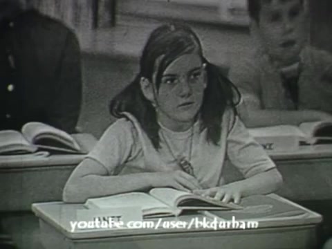 The still from '6th Grade Linguistics' (1966) features a young girl sitting at a desk, focused on an open textbook. Her hair is styled in pigtails, and she appears engaged in the classroom environment. The monochromatic aesthetic resembles classic kinescope recordings, typical of educational films of that era. The background hints at a traditional classroom setting, contributing to the film's pedagogical context.