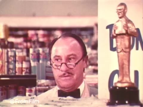 The still from the 'Charmin Commercial With Mr. Whipple' features a store setting with Mr. Whipple, a character known for his humorous approach to promoting Charmin toilet paper. He wears a white apron and bow tie, looking concerned as he peers over a display, conveying a sense of urgency about customers squeezing the Charmin. Beside him, there's a statuette, adding a whimsical touch to the scene. The vibrant color and graininess typical of a 16mm print capture the vintage charm of classic advertisements.   Follow us on Twitter @FromFilm.