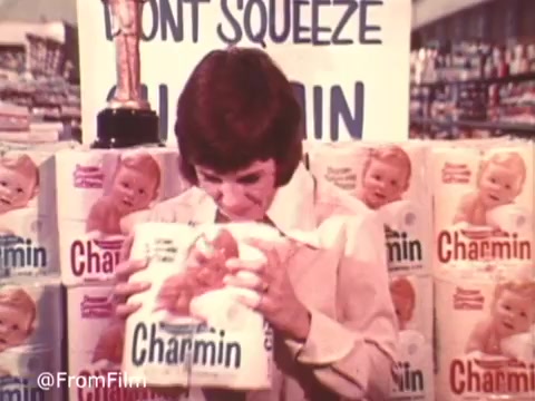 In this still from the 'Charmin Commercial With Mr. Whipple,' a woman is seen holding a package of Charmin toilet paper, expressing a comical moment of temptation as she prepares to squeeze it. She stands in front of a display of Charmin products, with a large sign reading 'Don't Squeeze the Charmin!' in the background. The nostalgic 16mm print captures the playful tone of the advertisement, emphasizing the product's softness.
