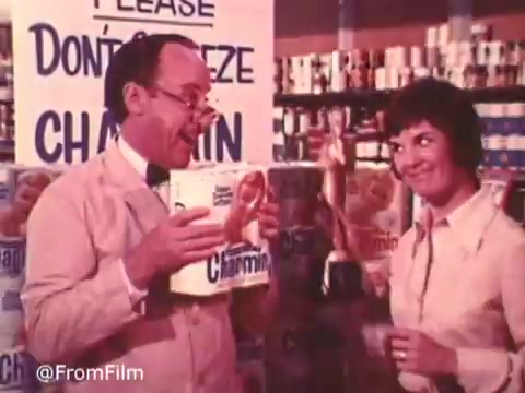 The still features a light-hearted scene from the iconic Charmin commercial with Mr. Whipple. Mr. Whipple, dressed in a suit, is animatedly promoting Charmin toilet paper, holding a box with a playful expression. A smiling woman stands nearby, seemingly amused by his antics. The backdrop displays shelves stocked with Charmin products, and a prominent sign reads 'Please Don't Squeeze the Charmin!' The vintage style highlights the charming, nostalgic feel of the ads from that era.   Follow us on Twitter @FromFilm.