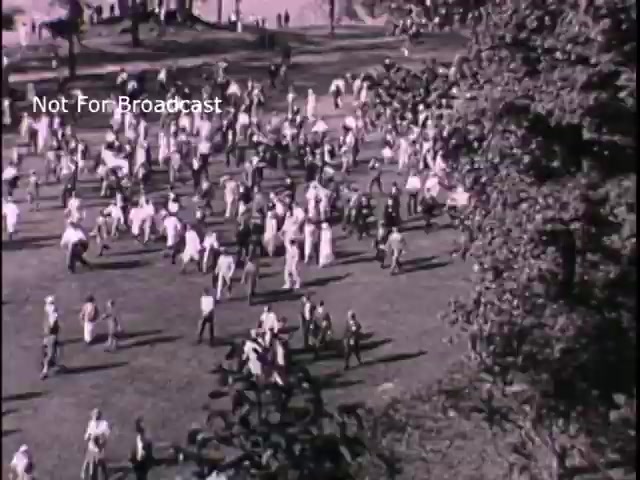 The still from the 1960s titled 'US Army recruiting - 'Join the new action army'' depicts a bustling outdoor gathering, likely aimed at highlighting the vibrancy and appeal of military service. The scene shows numerous people, possibly in a park setting, engaged in various activities, suggesting a community-focused event designed to attract potential recruits. The black and white imagery creates a nostalgic atmosphere, reflecting the era's recruitment efforts.