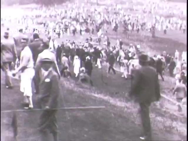 The still appears to depict a large crowd of people gathered outdoors, likely during a public event or rally promoting military recruitment for the U.S. Army in the 1960s. The scene captures a diverse group of individuals engaged in various activities, set against a backdrop of rolling hills or fields, evoking a sense of community and participation. The black-and-white format reinforces the historical context of the era.