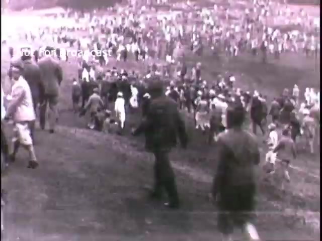 The still appears to show a large crowd of people gathered outdoors, possibly for a recruitment event or community gathering related to the U.S. Army in the 1960s. The scene is in black and white, capturing the era's aesthetic, with individuals dressed in period-appropriate attire. The atmosphere suggests a call to action, consistent with military recruitment themes.