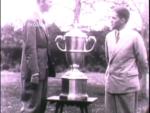 The 16mm still likely features two men in formal attire standing beside a trophy on a table, set in an outdoor environment. The focus appears to be on the trophy, symbolizing achievement or competition, possibly related to military recruiting efforts in the 1960s. The context suggests an attempt to attract recruits by associating military service with honor and accolades.