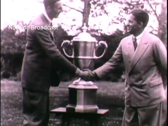 The still likely depicts two men shaking hands in front of a trophy. The setting appears to be an outdoor location, possibly a garden or park. The scene emphasizes camaraderie and achievement, aligning with themes of honor and teamwork that could resonate with the recruitment message of the 1960s U.S. Army. The visual suggests a celebration of success, potentially appealing to the audience's sense of duty and pride.