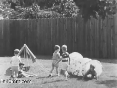 The still from the 'March of Dimes Birth Defects PSA' features a group of children playing outdoors. They're engaged in various activities, such as climbing on a small structure and playing around a circular play tunnel. The setting is a grassy area with a wooden fence in the background, suggesting a safe and playful environment. The image captures the innocence of childhood amidst the context of promoting awareness about birth defects.