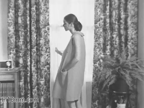 The still from the 'March of Dimes Birth Defects PSA' features a woman in a simple dress standing near a window with patterned curtains. She has a contemplative expression and appears to be looking out. The setting includes a radio-like device and a potted plant, suggesting a domestic atmosphere. This moment likely conveys a sense of reflection or concern related to the topic of birth defects and child health.