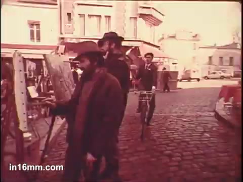 The still from the 'Birdseye Frozen Parisian Vegetables' commercial showcases a quaint Parisian street scene. It features several characters: a man with a hat and beard walking past an easel, alongside another individual on a bicycle. The atmosphere has a nostalgic, cinematic quality, enhancing the charm of the setting, which is characterized by cobblestone streets and classic architecture. The warm colors evoke a sense of homeliness and rustic appeal, aligning with the theme of fresh, quality vegetables associated with the brand.