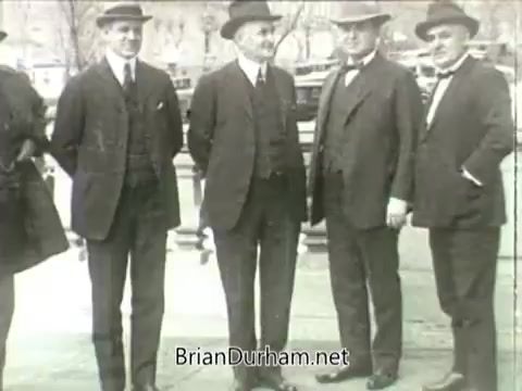 The still features a group of five well-dressed men standing together outdoors, likely from the early 20th century. They are all wearing hats and formal attire, indicative of the era. The background appears somewhat blurred, emphasizing the figures in the foreground. This scene is a part of newsreel footage related to John L. Lewis and the 1919 Miners' Strike, capturing a moment of organized labor history. The lack of intertitles means the specific context or actions are not provided, relying on visual cues alone.