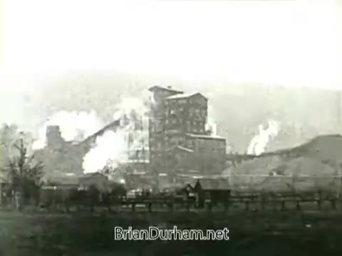 The still from 'Newsreel footage: John L. Lewis and the 1919 Miners' Strike' depicts a coal mine site with significant industrial activity. Smoke billows from the structures, indicative of mining operations, while the foreground features a wooden fence and a small building, suggesting a rural setting. The image captures the era's industrial landscape and the tensions surrounding labor disputes during that period.