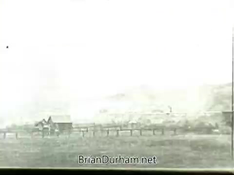 The still from 'Newsreel footage: John L. Lewis and the 1919 Miners' Strike' depicts a landscape with simple structures and a fence, possibly representing a mining town. The image appears faded and lacks clarity, characteristic of older film footage. This stark visual captures the socioeconomic context of the miners' strike era, emphasizing the harshness of labor conditions.