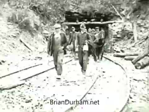 The still depicts a group of miners walking along a curved track leading into a coal mine entrance. They are dressed in work clothing typical of the early 20th century, and the environment appears rugged, surrounded by rocky terrain and vegetation. The image captures the hard labor and camaraderie of the miners during the 1919 Miners' Strike, reflecting the historical context of labor disputes in the coal mining industry.