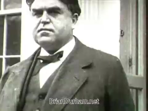 The still from the 16mm newsreel footage features John L. Lewis, a significant figure in labor history, prominently displaying a serious expression. He is dressed in a formal suit with a bow tie, suggesting an important public role or statement. The background is blurred yet appears to include a building, indicating a setting relevant to the miners' strike of 1919. The overall tone conveys the weight of the labor issues at the time.