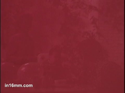 The still appears to be heavily tinted in red, obscuring details. There seem to be figures in the background, possibly adults and a child, suggesting a domestic setting. The overall composition hints at a focus on family and the act of feeding, characteristic of baby food commercials. The visibility is limited due to the color distortion.