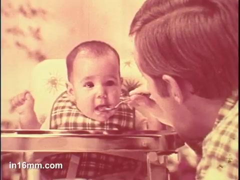 The still features a baby in a high chair, eagerly reacting to being fed by an adult. The baby appears to be enjoying the food, with a cheerful expression. The setting has a warm, home-like ambiance, and the colors are soft and nostalgic, typical of vintage commercials. The scene captures a tender moment between the adult and the child, emphasizing the product's appeal for parents.