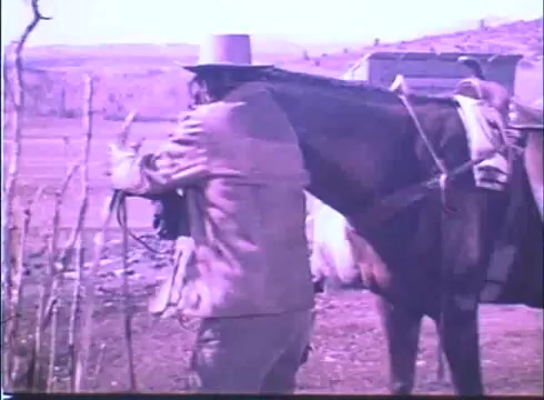 The still from the 'Showdown' trailer features a cowboy in a light-colored suit and hat, standing beside a horse. He is holding onto the horse's reins, set against a backdrop of a rugged landscape. The color tone appears faded, typical of a 16mm print, lending a vintage feel to the scene. The overall atmosphere suggests a western theme.